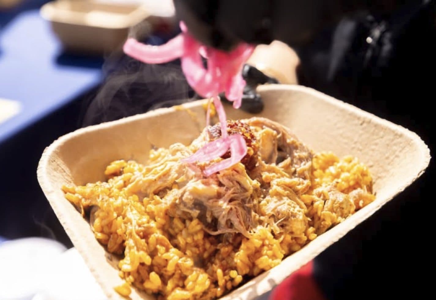Close-up of a compostable bowl of paella being topped with bright pickled onions