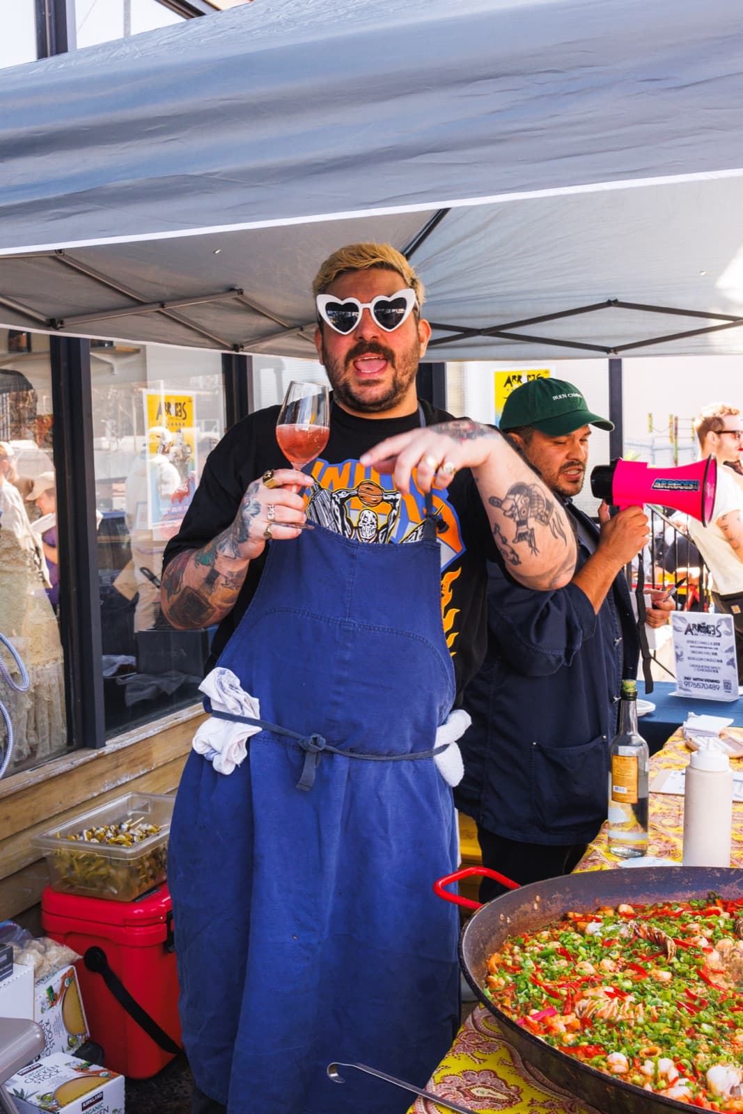 Chef in heart-shaped sunglasses holding a glass beside a full paella pan