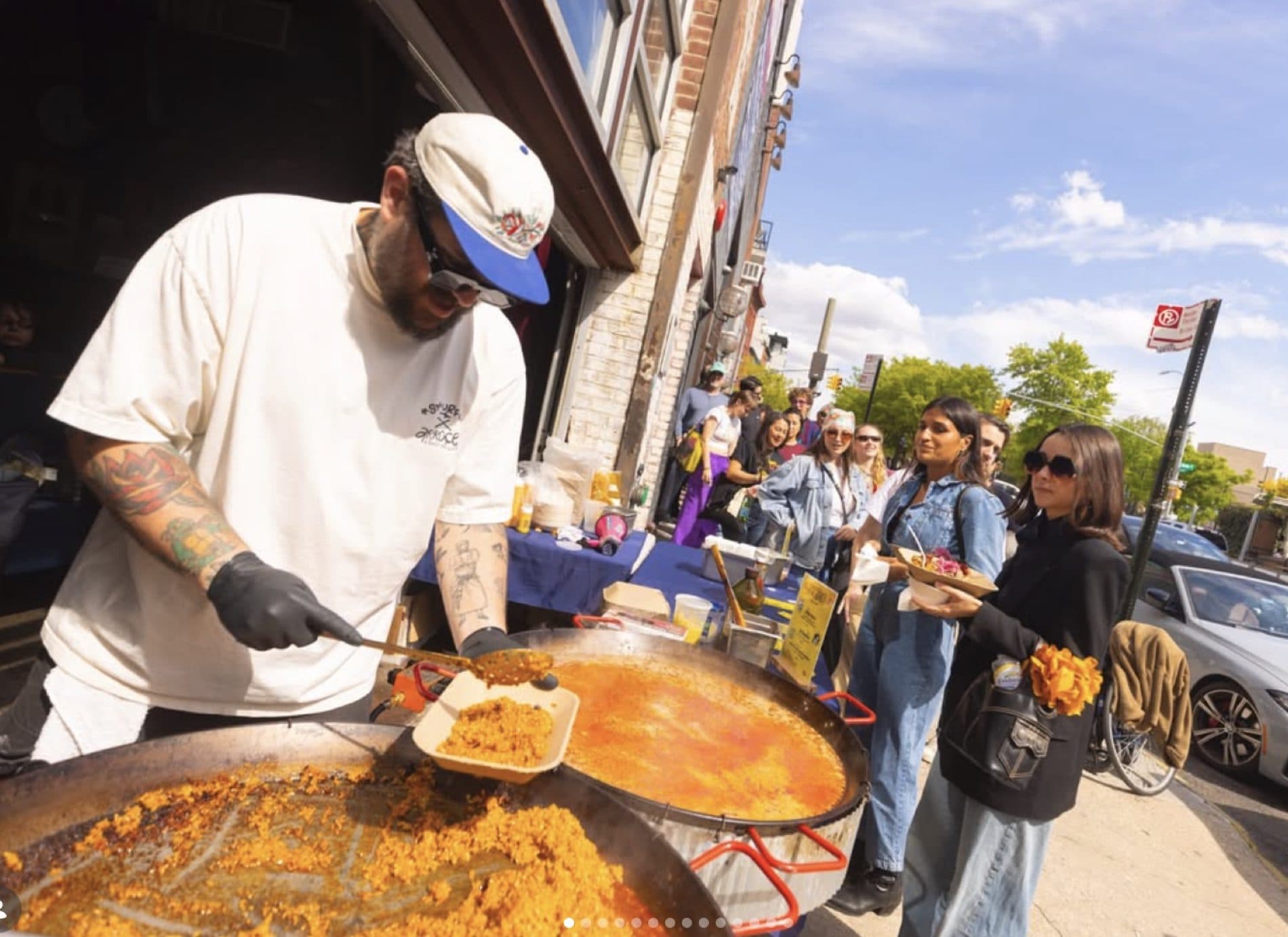 Chef Eduardo serving paella from large pans to a line of guests on a sunny New York sidewalk