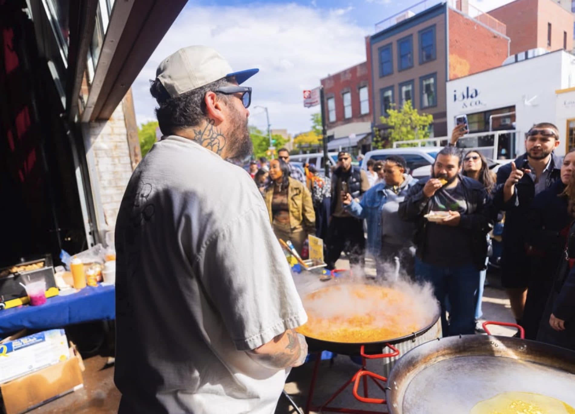 Chef Eduardo Cuenca stirring a large steaming paella pan at an outdoor New York pop-up while guests look on